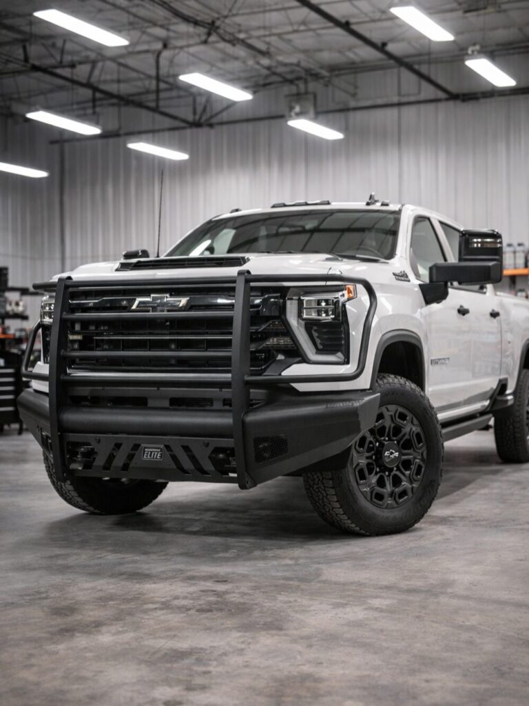 White heavy-duty pickup truck with black steel grille guard and elite front bumper installed, photographed inside a clean, professional truck accessories shop with polished concrete floors and overhead lighting.